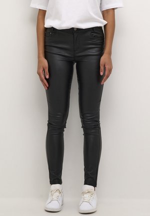 Person trägt schwarze Lederhose, weißes T-Shirt und weiße Sneakers und steht vor einem schlichten hellen Hintergrund.