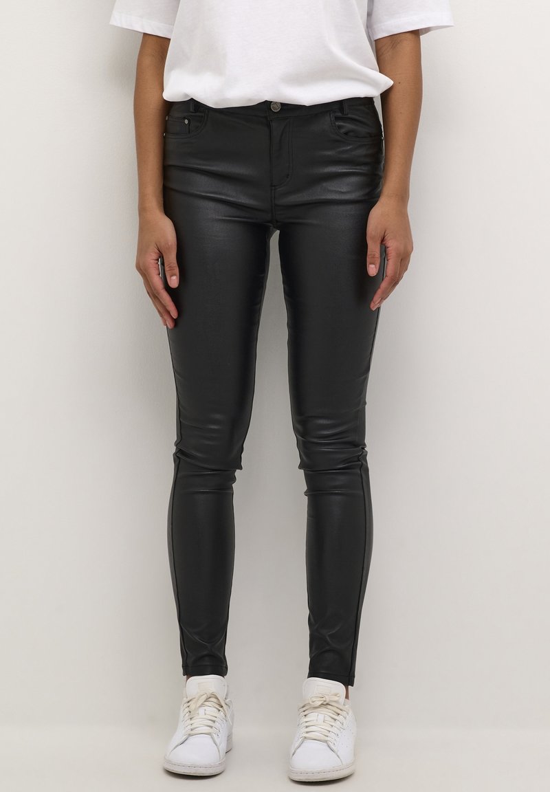 Personne portant un pantalon en cuir noir, un t-shirt blanc et des baskets blanches, debout devant un fond clair uni.