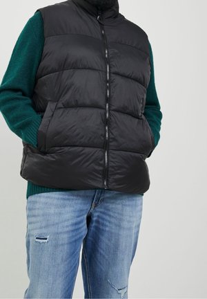 Mężczyzna w czarnej pikowanej kamizelce na zielonym swetrze, z rękami w kieszeniach, w jasnoniebieskich, podartych dżinsach na szarym tle.