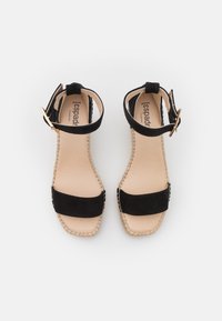 Black suede sandals with ankle straps, featuring a flat espadrille sole, beige footbed, and natural stitching along the edges.