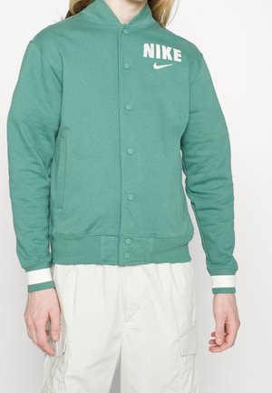 Osoba nosząca zieloną kurtkę Nike na guziki z białym logo na piersi i białe spodnie typu cargo na jednolitym tle.