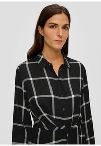 Schwarz-weiße karierte Bluse mit langen Ärmeln, Knopfverschluss vorne und einem gebundenen Taillendetail. Texturiertes Material mit sichtbarem Gittermuster.