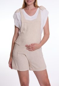 Beige linnen short overalls met verstelbare schouderbanden, een voorzak en omgeslagen zoom, gedragen over een witte V-hals t-shirt.