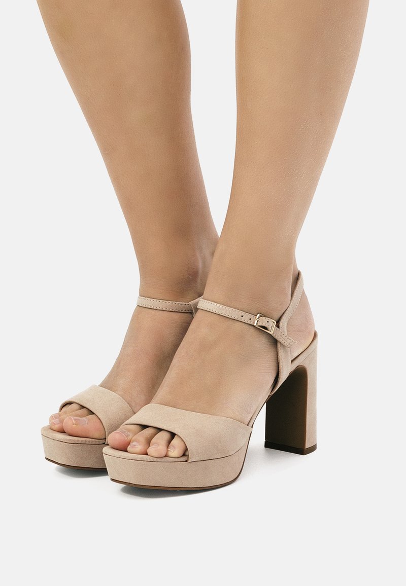 Sandalias de tacón alto de ante beige con plataforma gruesa, punta abierta y correa de tobillo con cierre de hebilla. Textura suave, diseño minimalista.