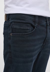 Prim-plan cu buzunarul blugilor bleu închis care ține un smartphone negru, purtat cu o cămașă albă băgată în pantaloni.