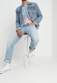 Chaqueta de denim azul claro sobre una camiseta rosa, combinada con jeans azul claro y zapatillas blancas. Sentado en un taburete blanco frente a un fondo blanco.