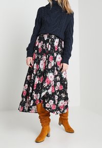 Nainen, joka on pukeutunut tummansiniiseen palmikkoneuleeseen, mustaan kukkakuosisesti midi-hameeseen, jossa on vaaleanpunaisia kukkia, ja rentoihin sinapinkeltaisiin mokkabootseihin, joissa on paksut korot.