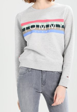 Kobieta ubrana w jasnoszarą bluzę Tommy Jeans z kolorowymi paskami oraz dopasowane niebiesko-szare dżinsy, stoi z jedną ręką rozluźnioną.