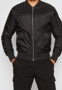 Svart bomberjacka i glänsande material, med dragkedja framtill, ribbstickade muddar och två sidofickor samt en användbar ficka på ena ärmen.