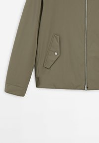Section de veste vert olive avec manche longue, fermeture éclair argentée et poche inclinée avec rabat et bouton-pression sur fond blanc.
