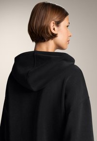 Felpa nera con cappuccio, dal taglio rilassato, cuciture visibili sui bordi e una texture morbida. Presenta una tasca a marsupio sul davanti.