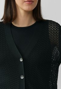 Schwarze Strickjacke mit V-Ausschnitt, strukturiertem Zickzackmuster und glatten Knöpfen. Kombiniert mit einem schwarzen, gerippten Oberteil darunter.