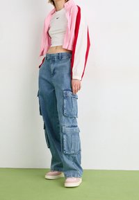 Chaqueta corta de color rosa y blanco con cremallera, camiseta blanca ajustada, pantalones cargo azules holgados con grandes bolsillos y zapatillas con suela rosa.