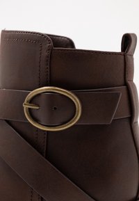 Bota de tobillo de piel marrón con un hebilla de latón redonda y diseño de correas cruzadas. Textura suave con detalle de costura a lo largo de los bordes.