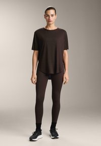 Brązowa koszulka z krótkim rękawem o luźnym kroju, zestawiona z dopasowanymi legginsami i czarnymi butami sportowymi. Gładki materiał z minimalnym wzorem.