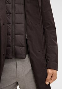Donkerbruine, gestructureerde buitenjas over een gewatteerde binnenlaag. Voorzien van een ritssluiting en zijzakken. Lichte grijze op maat gemaakte broek zichtbaar.