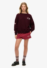 Sudadera burdeos con el texto "NEW YORK", de corte holgado y puños de canalé, combinada con pantalones cortos de color bayas a juego y calcetas de patrón, y zapatos negros.