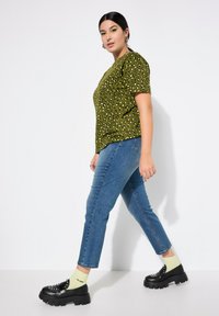 Camiseta de leopardo verde, jeans ajustados azules y zapatos negros gruesos. Presenta mangas cortas y un corte relajado. Medias amarillas visibles.