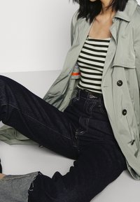 Vaaleanvihreä trench-takki, jossa on kaksi suurta nappia, päällä musta-valkoraidallinen toppi, yhdistettynä tummansinisiin korkeavyötäröisiin farkkuihin.