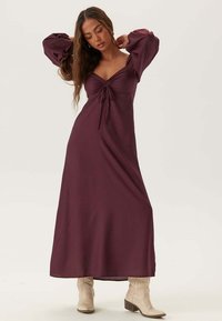 Lungo abito bordeaux con maniche a sbuffo, scollo a cuore e dettagli con stringa sul busto. Indossato con stivali cowboy beige.
