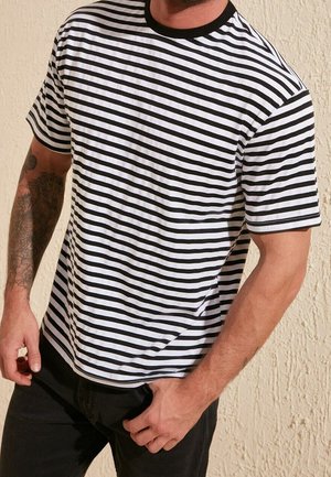 Mann trägt kurzärmeliges schwarz-weiß horizontal gestreiftes T-Shirt mit schwarzem Kragen, kombiniert mit schwarzer Hose, steht vor einer strukturierten Wand.
