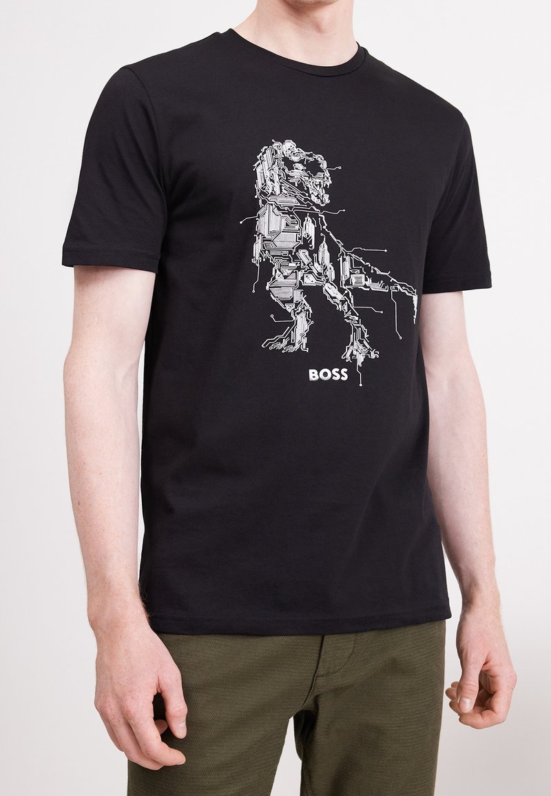 Svart t-shirt med vit dinosaurgrafik i kretskortsstil och texten "BOSS" som bärs av en person i olivgröna byxor.