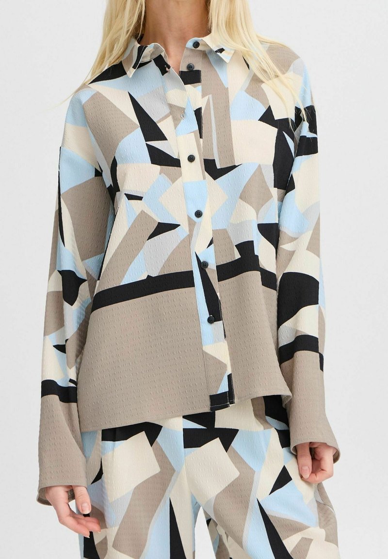 Femme portant une chemise ample beige, bleue, noire et blanche à motif géométrique, boutonnée, avec un pantalon assorti.