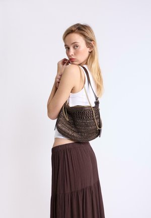 Lyshåret kvinde i hvid tanktop og brun nederdel bærer en tekstureret mørkebrun håndtaske med guldkæde-remme over skulderen.