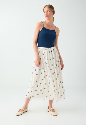 Femme portant un haut sans manches bleu marine et une jupe midi blanche à pois bleu marine, chaussures plates beiges, debout devant un fond blanc uni.