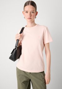 T-shirt di cotone rosa chiaro con colletto tondo e maniche corte, con un piccolo logo ricamato. Abbinato a pantaloni verde oliva e una borsa nera.