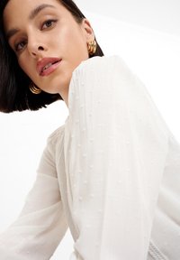 Blusa blanca texturizada con patrón de puntos, que muestra una tela delicada y mangas abullonadas. Pendientes de aro dorados complementan el conjunto.