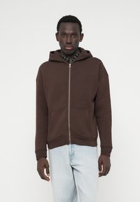 Fiatal férfi sötétbarna zipzáros kapucnis pulóverben, amely alatt kockás ing van, világoskék farmernadrággal áll egy sima fehér háttér előtt.