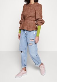 Femme portant un pull marron en maille avec des manches bouffantes, un haut vert à manches longues en dessous, un jean en denim clair déchiré et des baskets roses.