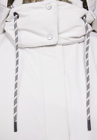 Vit jacka med hög krage, utrustad med svart- och vitdragkedjor, silverfärgade tryckknappar och en slät, texturerad tygfinish.