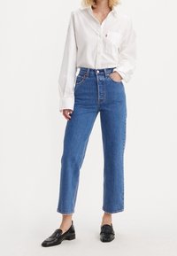 Vit skjorta med långa ärmar, blå högmidjade jeans med raka ben och svarta slip-on skor. Jeans har en klassisk femfickdesign.