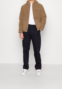 Felpa con zip di colore marrone, in pile, con una superficie testurizzata, abbinata a pantaloni blu navy e scarpe da ginnastica bianche.