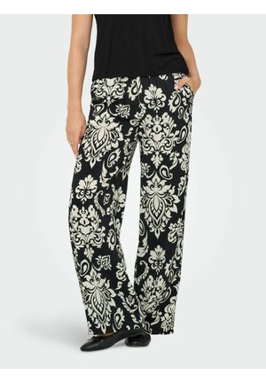 Femme portant un pantalon large noir et blanc avec un motif floral damassé complexe, associé à un haut noir et des chaussures plates noires.