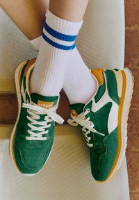 Baskets en daim vert avec des accents blancs, des détails orange et jaunes, des lacets blancs et des semelles texturées, assorties à des chaussettes blanches à rayures bleues.