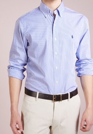 Homme portant une chemise à rayures bleu clair avec boutons et manches retroussées, pantalon beige et ceinture noire avec une boucle dorée.