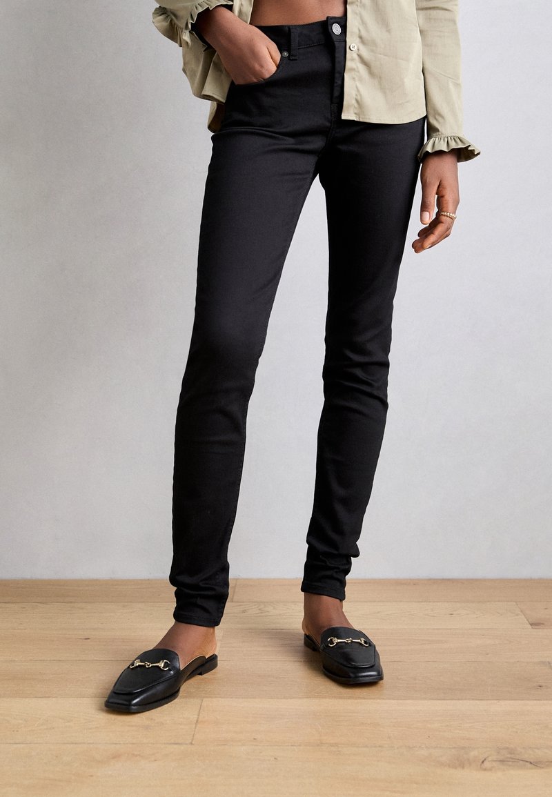 Persona vistiendo pantalones negros ajustados, mocasines negros sin cordones con acentos dorados y una camisa de manga larga beige con puños con volantes.