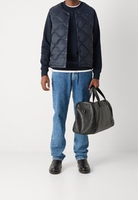 Schwarze Leder-Dufflebag mit runder Form und doppelten Griffen, getragen mit einer navyblauen gesteppte Weste über einem navyblauen Sweatshirt und hellblauen Jeans.