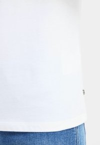 Hvid bomulds t-shirt med lige skåret kant, prydet med et lille mærke på venstre side, bæres med blå denimjeans.