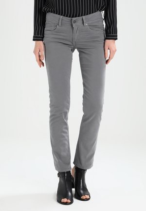 Frau trägt graue, schmal geschnittene Hose kombiniert mit schwarzen offenen Knöchel-Stiefeln und einem schwarzen Langarmshirt mit weißen Streifen.