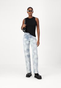 Top negro sin mangas, jeans celestes con un patrón de serpiente, zapatos de plataforma negros y gafas de sol; la modelo está de pie contra un fondo liso.
