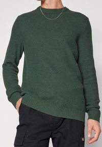 Dunkelgrüner Strickpullover mit rundem Ausschnitt, gerippten Bündchen und Saum sowie strukturiertem Stoff. Über dunklen Cargohosen getragen.