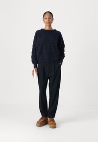 Maglione lavorato a maglia di colore navy con motivi testurizzati, abbinato a pantaloni neri larghi e scarpe con plateau marroni. La modella tiene una piccola borsa bianca.