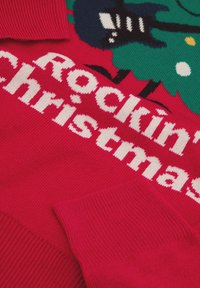 Maglione rosso lavorato a maglia con scritta bianca "Rockin' Christmas", con elementi grafici di un albero di Natale verde e una chitarra nera.