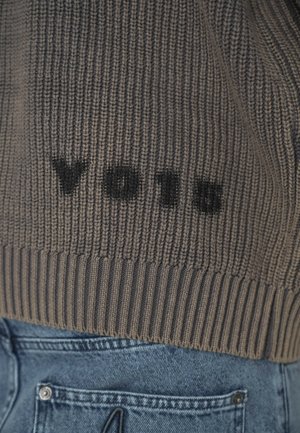 Hnedý pletený sveter s čiernym textom "VOIR" na zadnej kapsičke modrých džínsov.