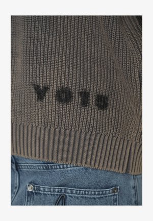 Suéter de punto marrón con texto negro "VOIR" sobre el bolsillo trasero de unos jeans de mezclilla azules.