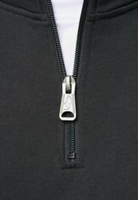 Crna zip-up odjeća s metalnim patentnim zatvaračem i logotipom na povlačnom dijelu. Tkanina izgleda glatko i rastezljivo, pogodna za svakodnevno nošenje.